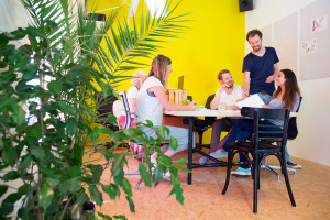 Designers, sitting in at a large table in a creative environment and office, surrounded by tack boards with drawings, plants and a bright yellow wall